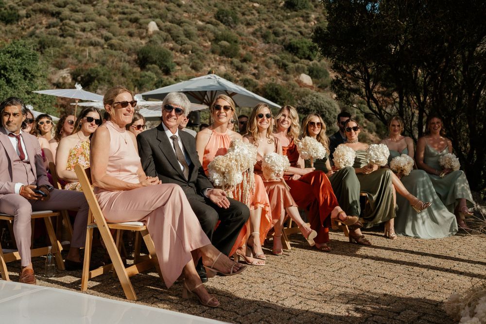 Wedding guests seated in wooden chairs at an outdoor ceremony against a hillside backdrop.