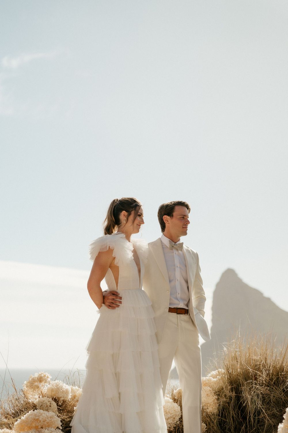 Newlyweds in white attire posing against a scenic mountain backdrop.