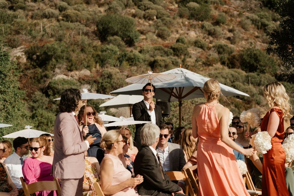 Wedding guests socialize at an outdoor reception under umbrellas with mountains in the background.