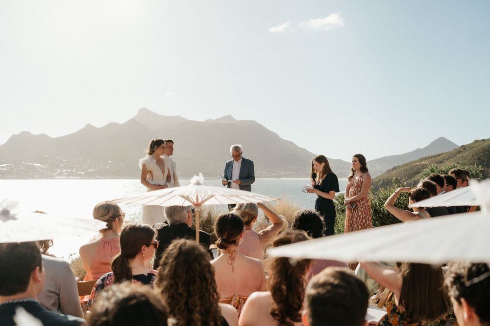 Outdoor wedding ceremony overlooking mountain ranges with guests seated under white umbrellas.