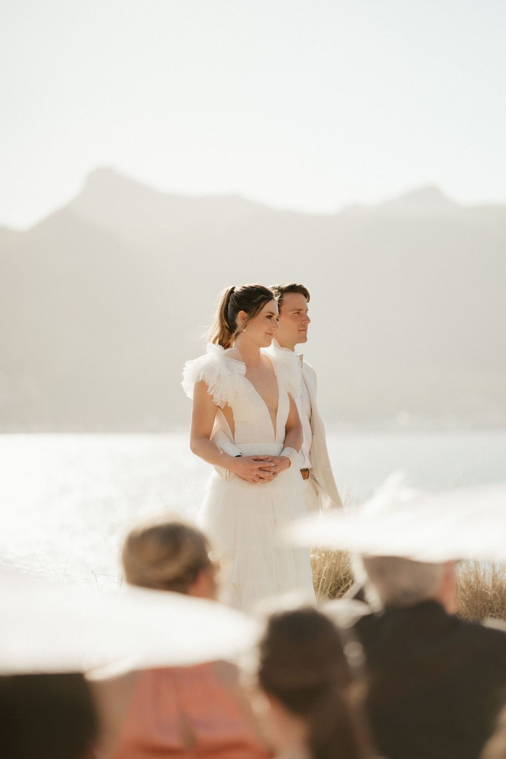 Couple in white wedding attire standing against mountain backdrop during outdoor ceremony.