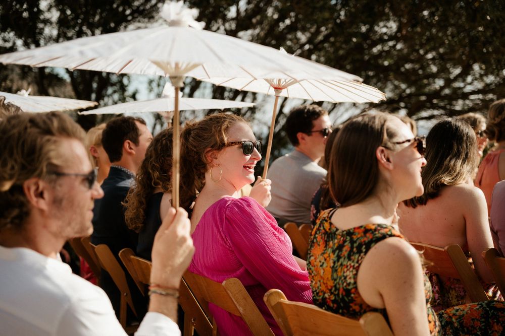 Outdoor wedding guests seated under white umbrellas during sunny ceremony.
