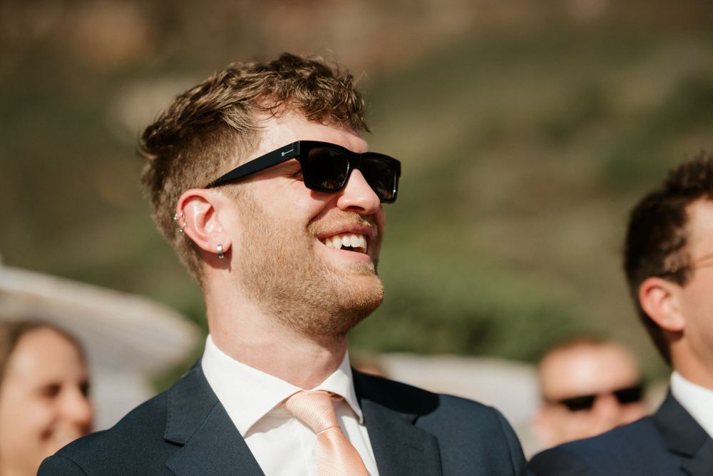 Smiling wedding guest wearing sunglasses and a suit with peach tie at an outdoor celebration.