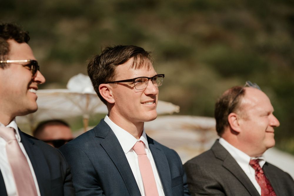 Wedding guests in formal attire smile during an outdoor ceremony.