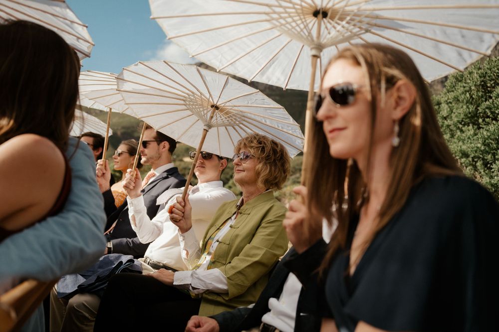 Wedding guests hold white parasols while enjoying an outdoor celebration on a sunny day.