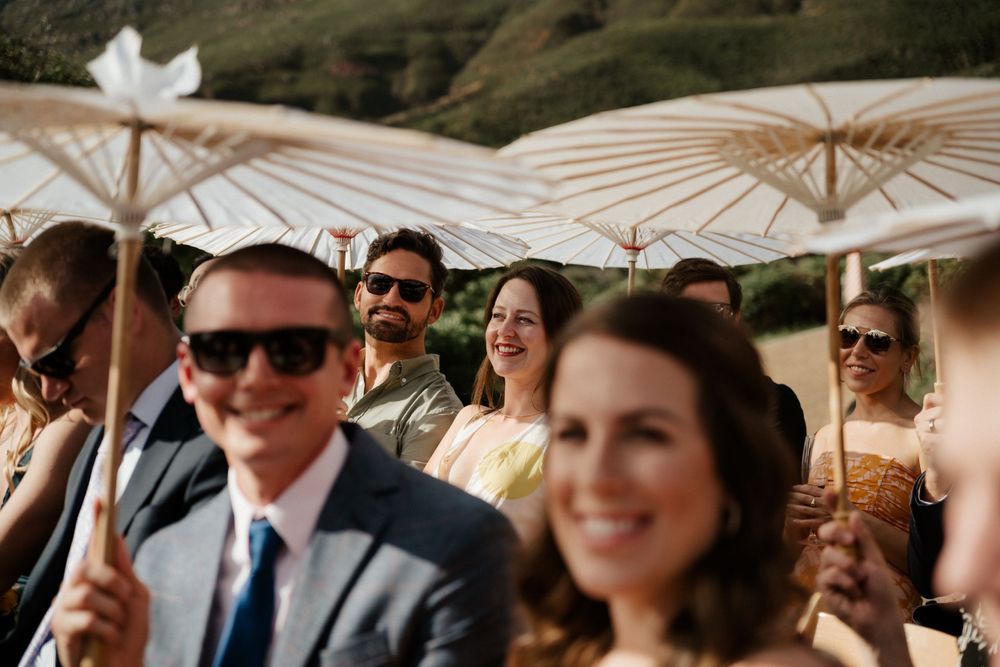 Wedding guests smile under elegant white paper parasols during an outdoor ceremony.