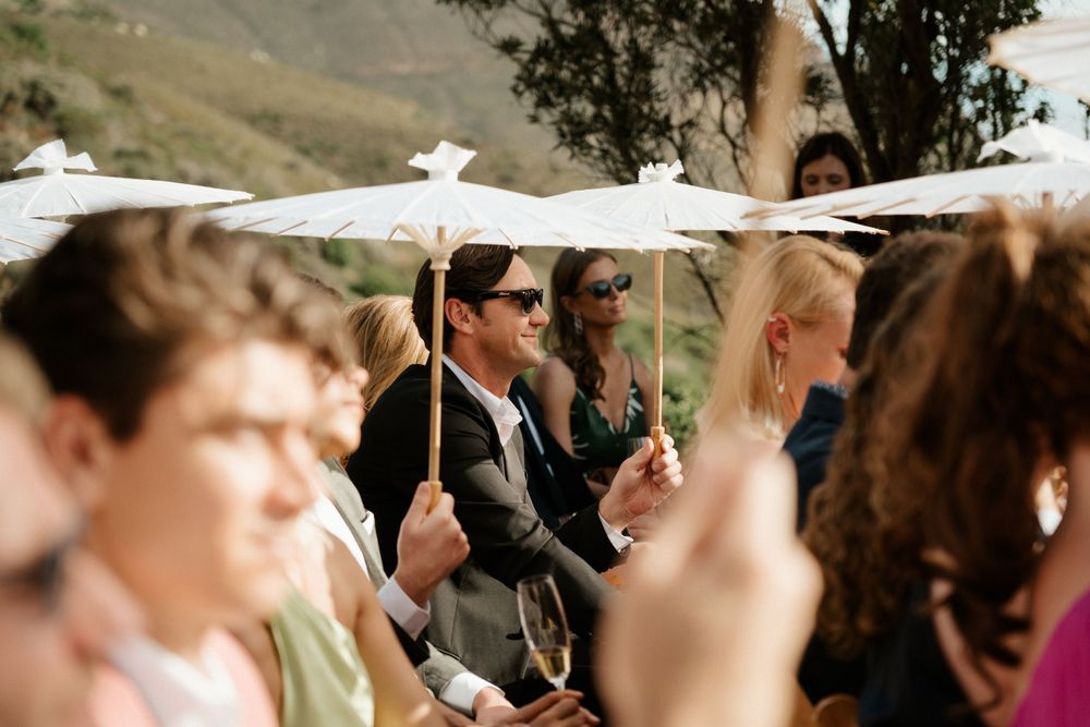 Wedding guests holding white parasols during outdoor mountain ceremony.