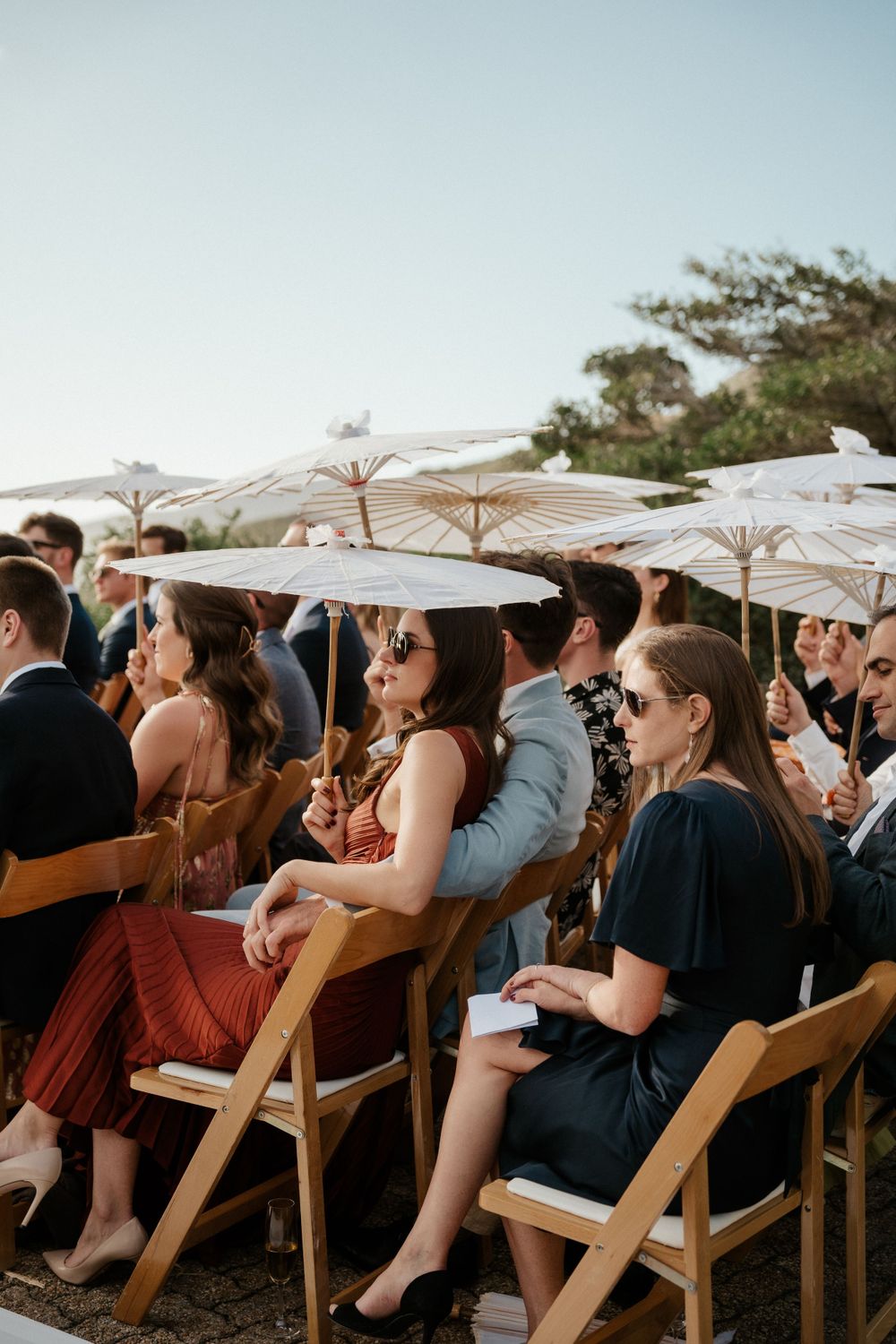 Wedding guests sit in wooden chairs holding white parasols during an outdoor ceremony.