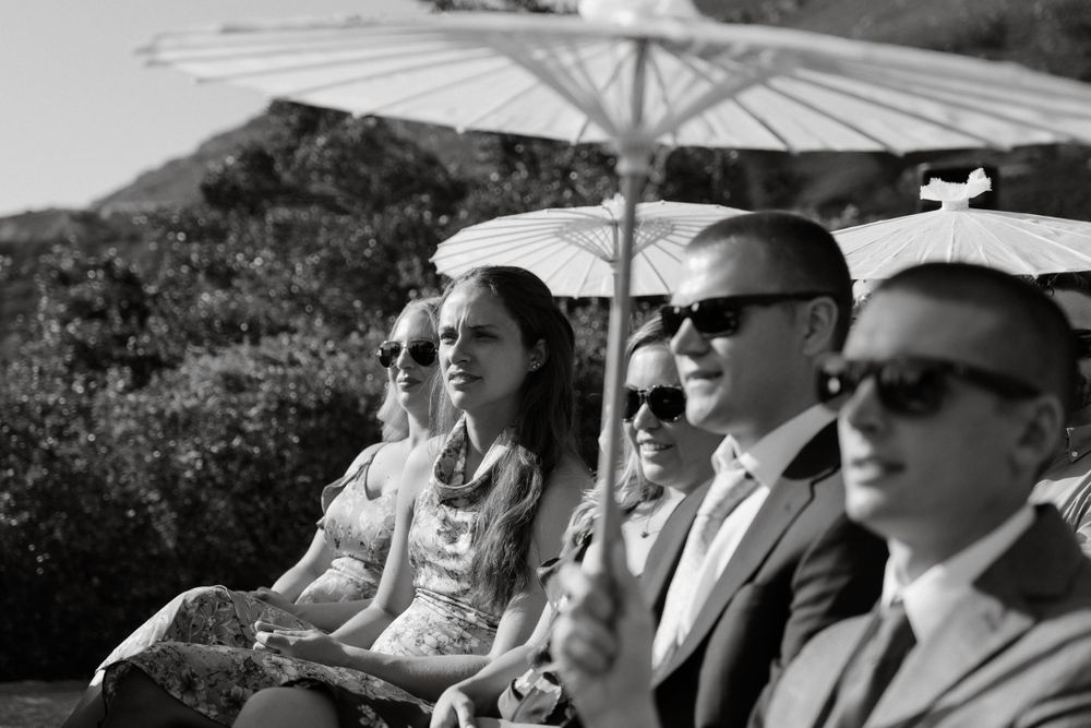 Black and white photo of wedding guests seated with parasols against mountain backdrop.