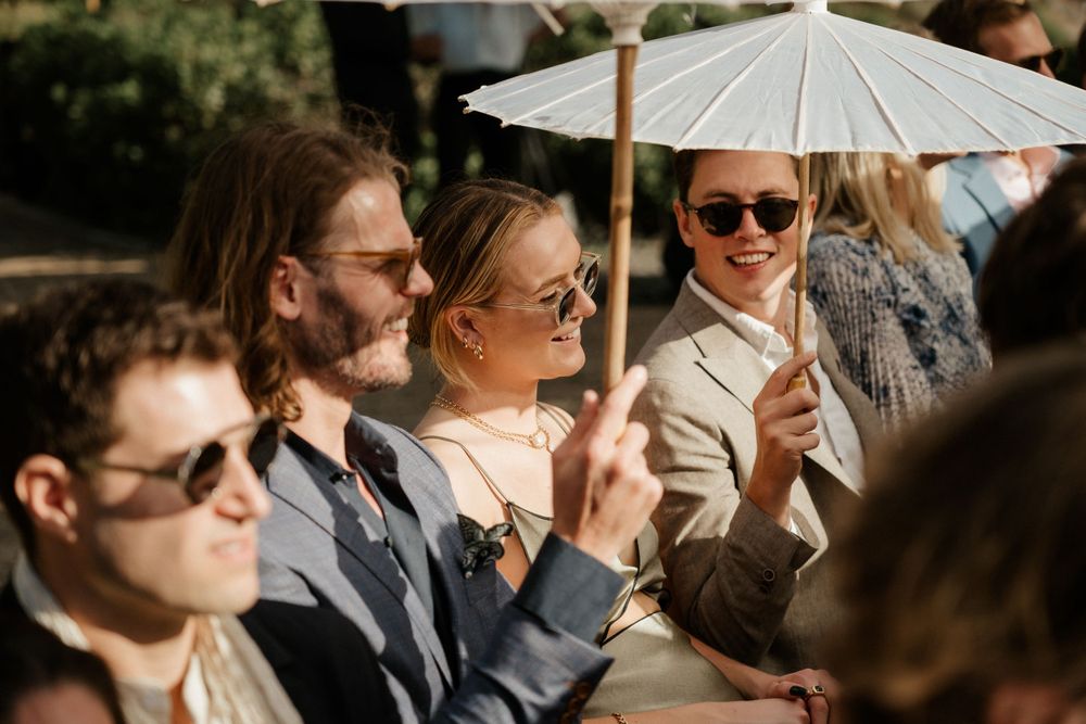 Wedding guests enjoying outdoor ceremony under white parasol umbrellas on sunny day.