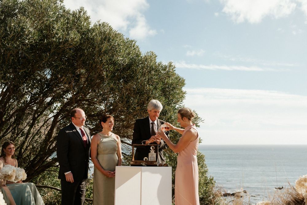 An intimate outdoor wedding ceremony takes place overlooking the ocean on a sunny day.