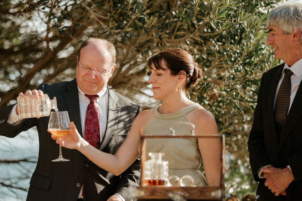 Wedding guests toasting with drinks during an outdoor celebration under olive trees.