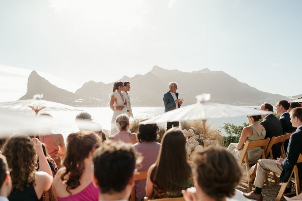Outdoor wedding ceremony with mountains and ocean views as guests watch the proceedings on the beach.