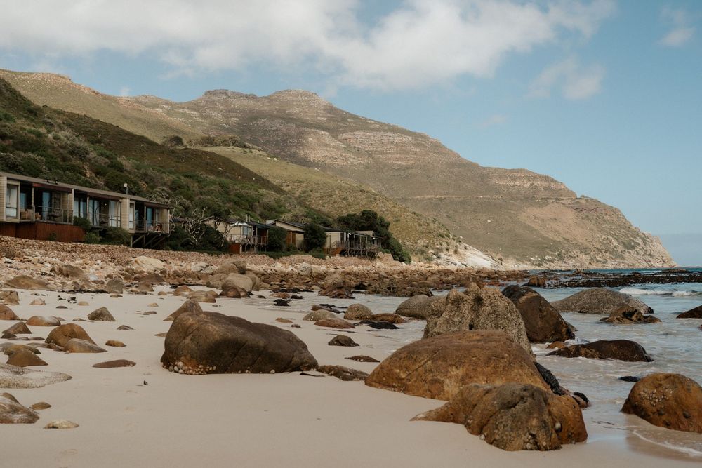 Coastal beach landscape with large rocks and mountains against blue sky.