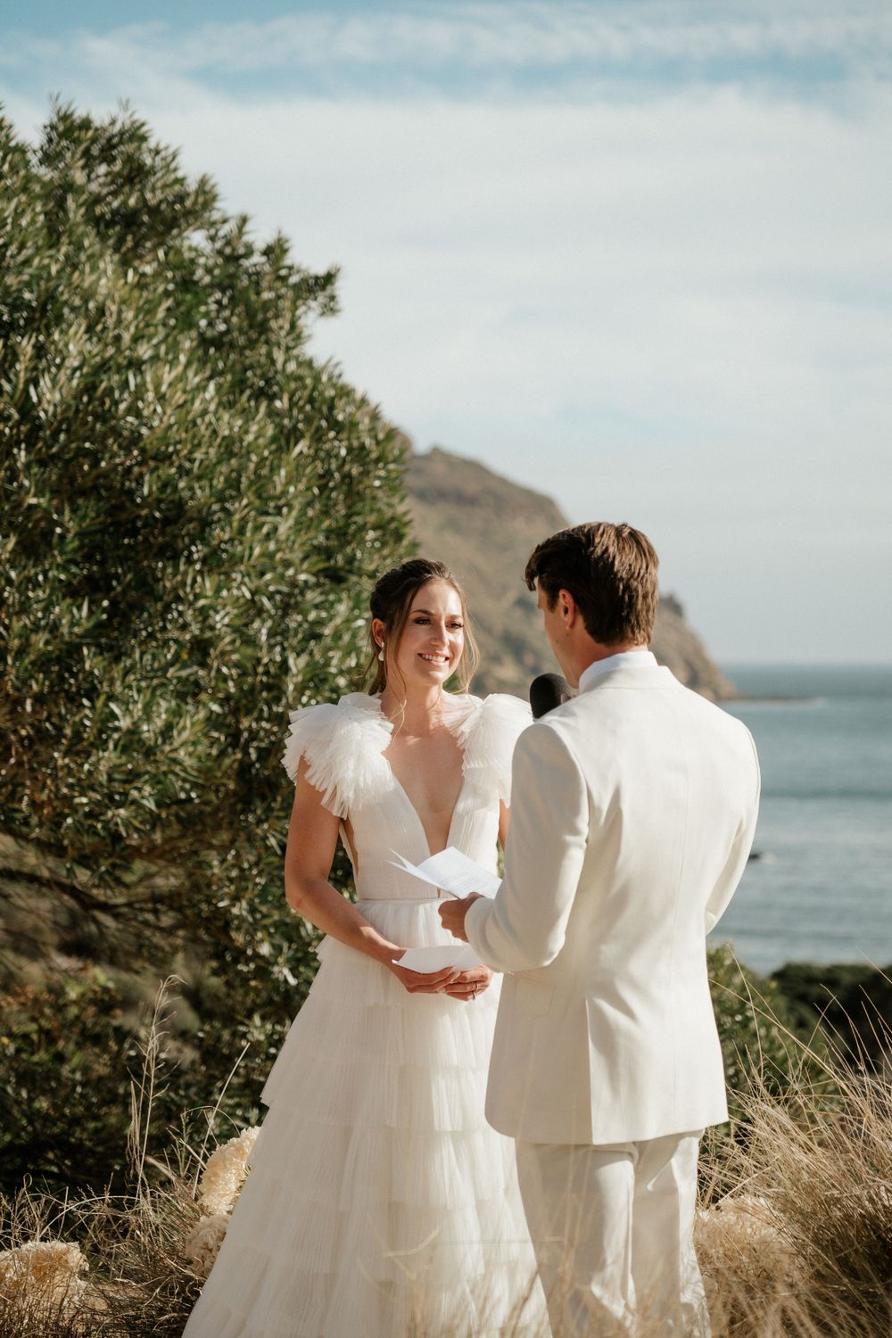 Couple in wedding attire standing together on coastal cliff overlooking ocean.