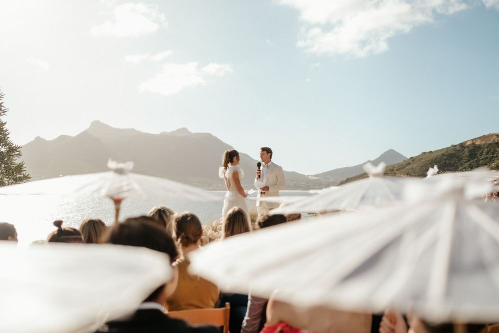 Outdoor mountain ceremony with white parasols and guests seated beneath a beautiful sunlit mountain landscape.