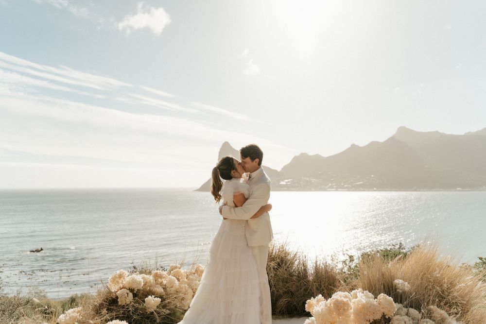 Newlyweds embrace on a coastal cliff with flowers and ocean views.