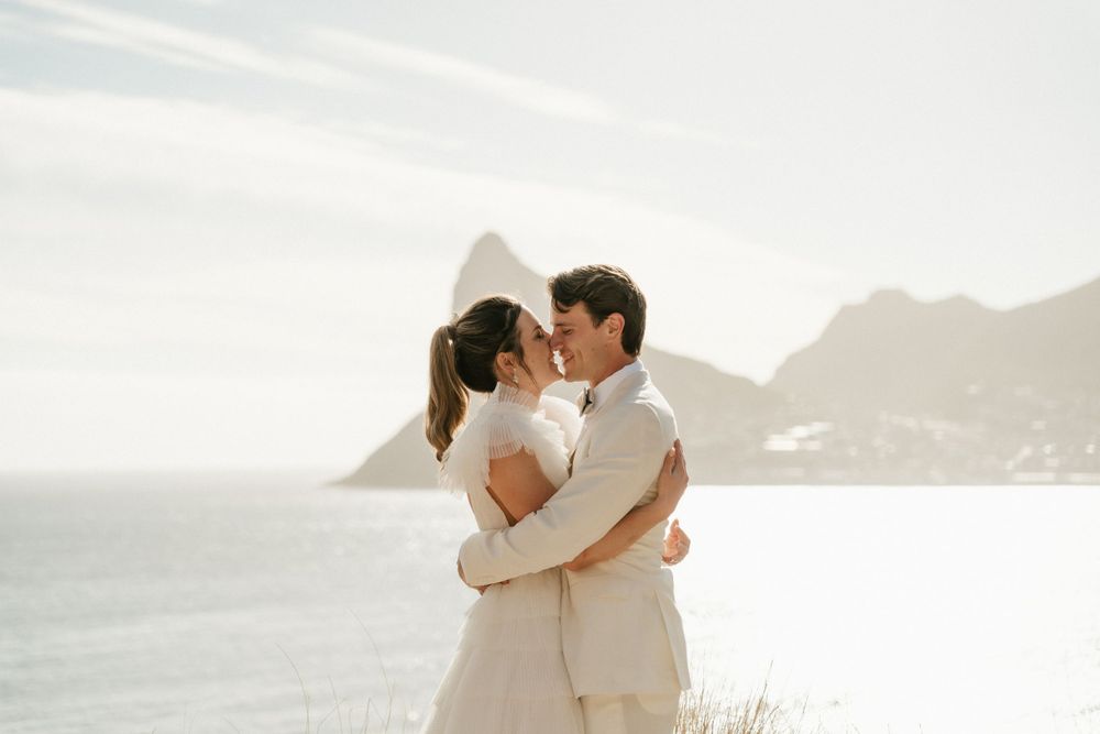 Newlyweds embrace with coastal mountain views in background during sunset.