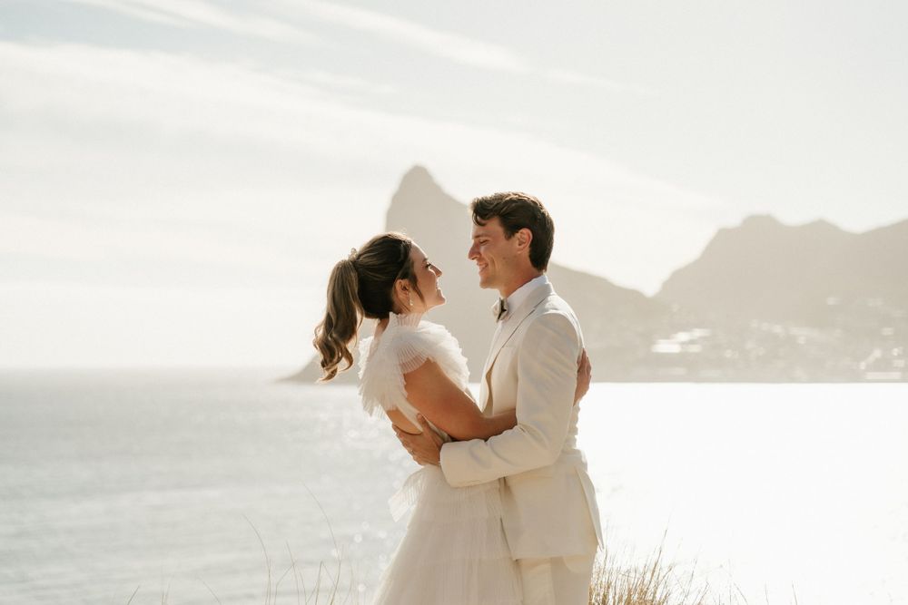 Romantic wedding couple embraces against dramatic coastal mountain backdrop at sunset.