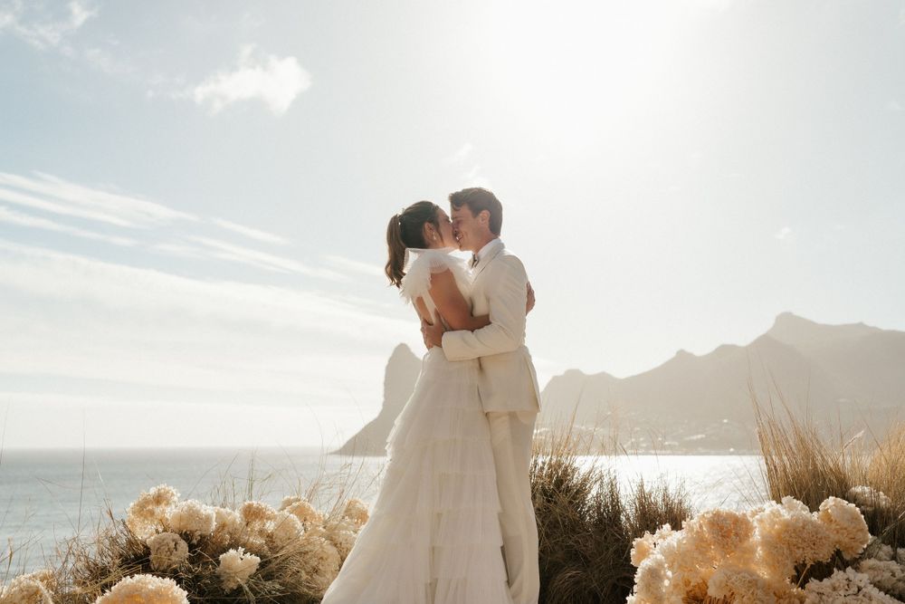 A couple embraces during their wedding ceremony overlooking ocean views with pampas grass decor at sunset.