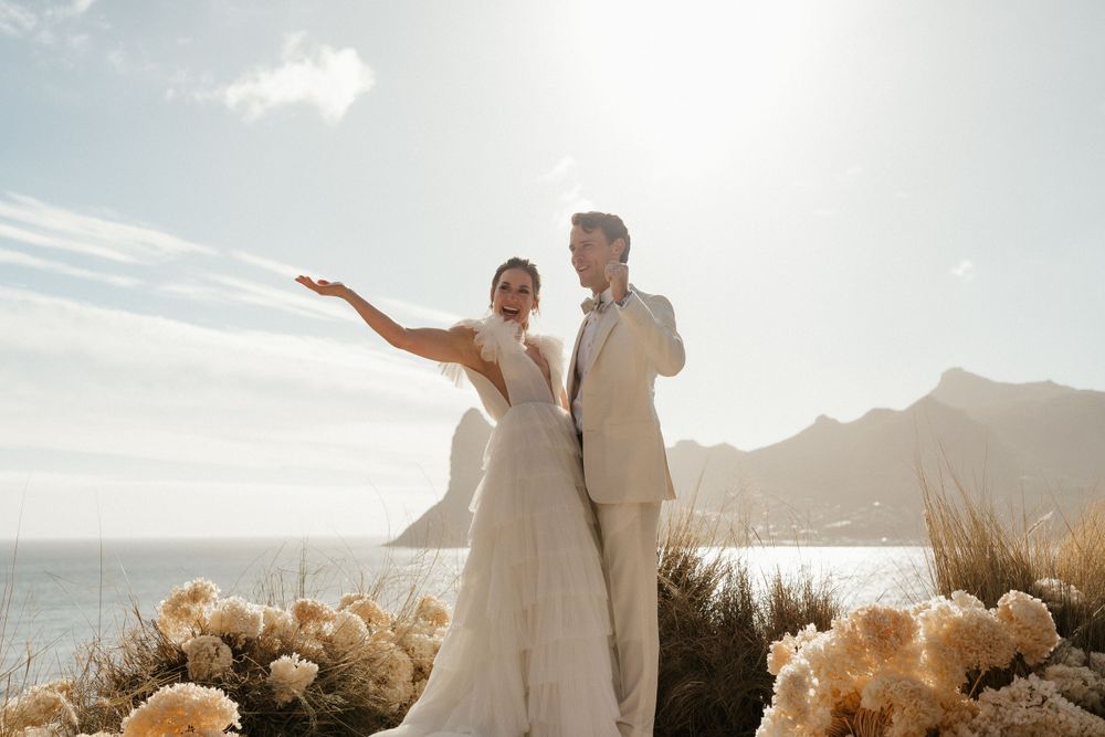 Wedding couple poses with dried pampas grass arrangements against ocean and mountain backdrop.