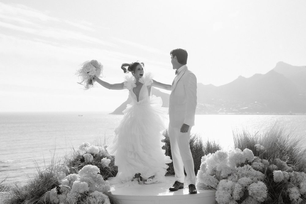 Black and white photo of couple dancing on coastal path with mountain backdrop.