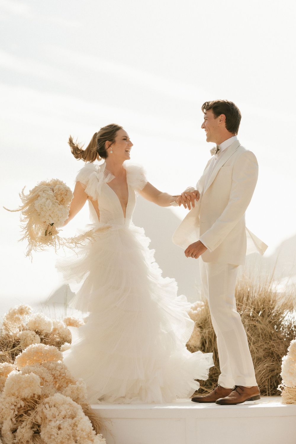Joyful wedding couple in all white attire dance together against scenic mountain backdrop.