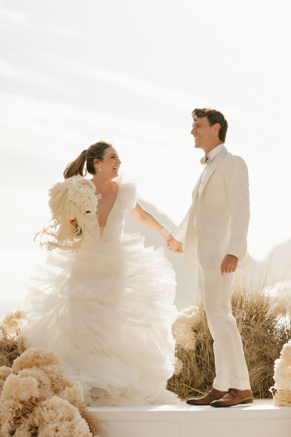 A wedding couple in white attire share a joyful moment outdoors with flowing fabric and pampas grass decorations.