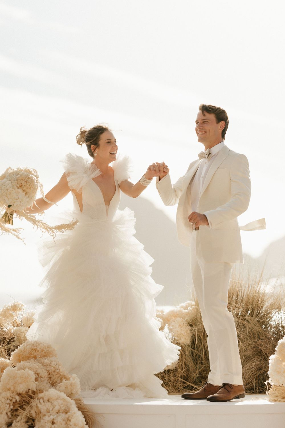 Couple in all-white wedding attire dance joyfully on a scenic outdoor platform.