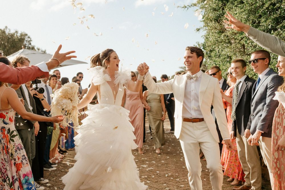 A wedding couple celebrates under confetti with guests throwing flower petals on a sunny outdoor path.