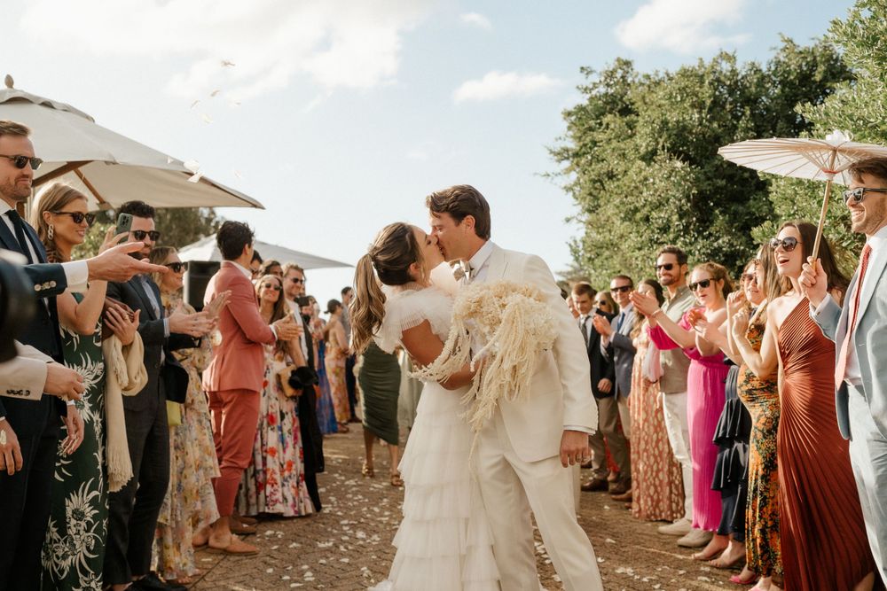 Wedding guests form an aisle outdoors while celebrating during a sunny outdoor ceremony.