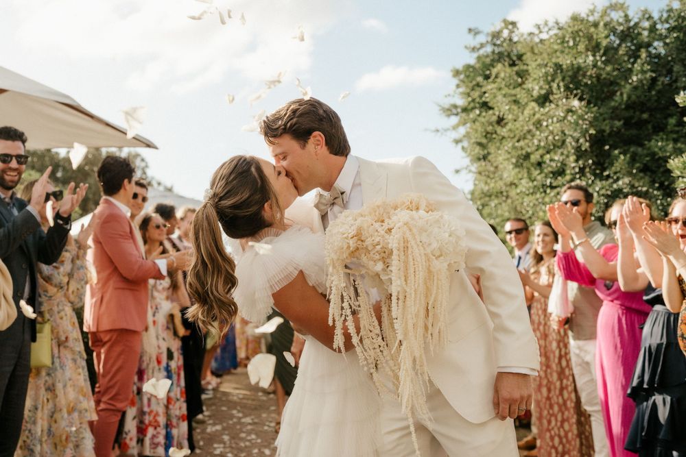 Newlyweds share romantic kiss during outdoor celebration as guests cheer with confetti.