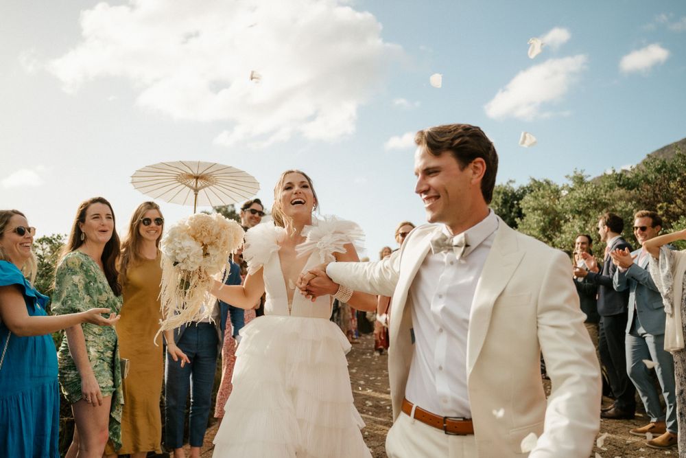Newlyweds celebrate under parasol surrounded by cheering wedding guests outdoors.