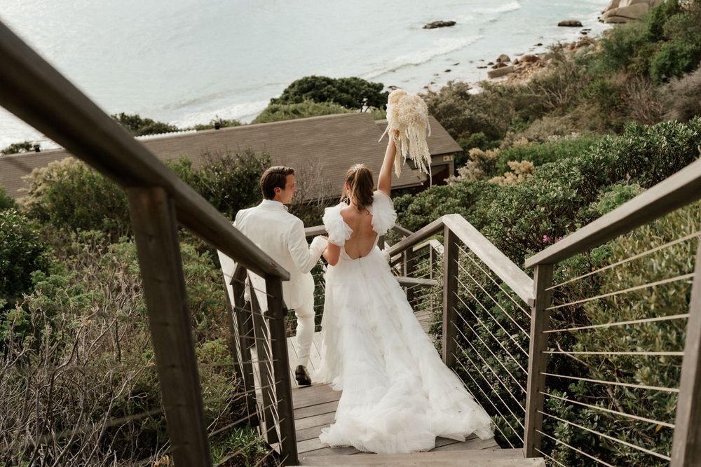 A couple descends outdoor stairs surrounded by coastal vegetation in wedding attire.