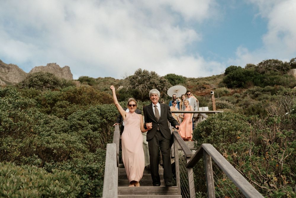 Wedding party celebrating on wooden stairs surrounded by lush coastal vegetation.