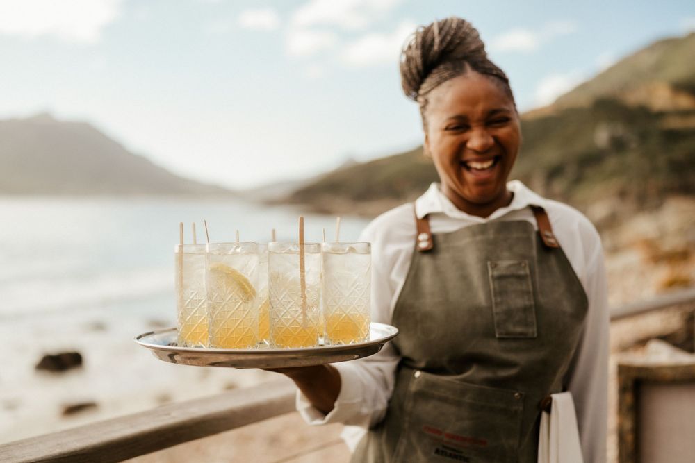 Server in apron holding tray of refreshing yellow cocktails against coastal backdrop.