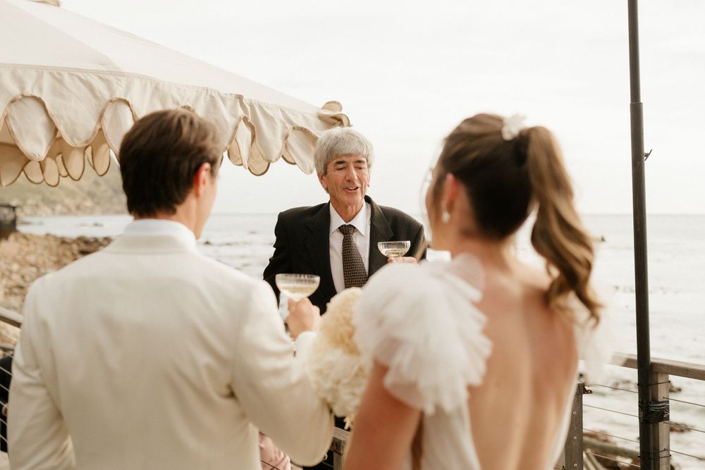 An intimate outdoor seaside wedding ceremony takes place under a beige canopy by the ocean.