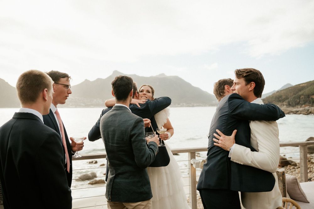 Wedding guests embrace and celebrate on oceanfront deck with mountain views.