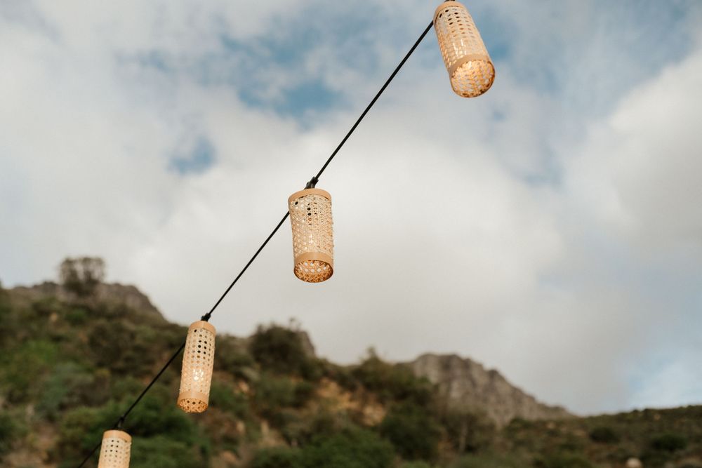 Rustic woven lanterns hang on strings against a mountainous backdrop with cloudy skies.