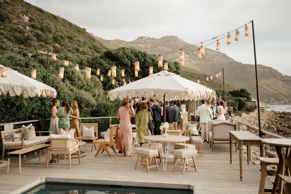 An outdoor wedding reception venue with white umbrellas and string lights on a mountainside terrace at sunset.