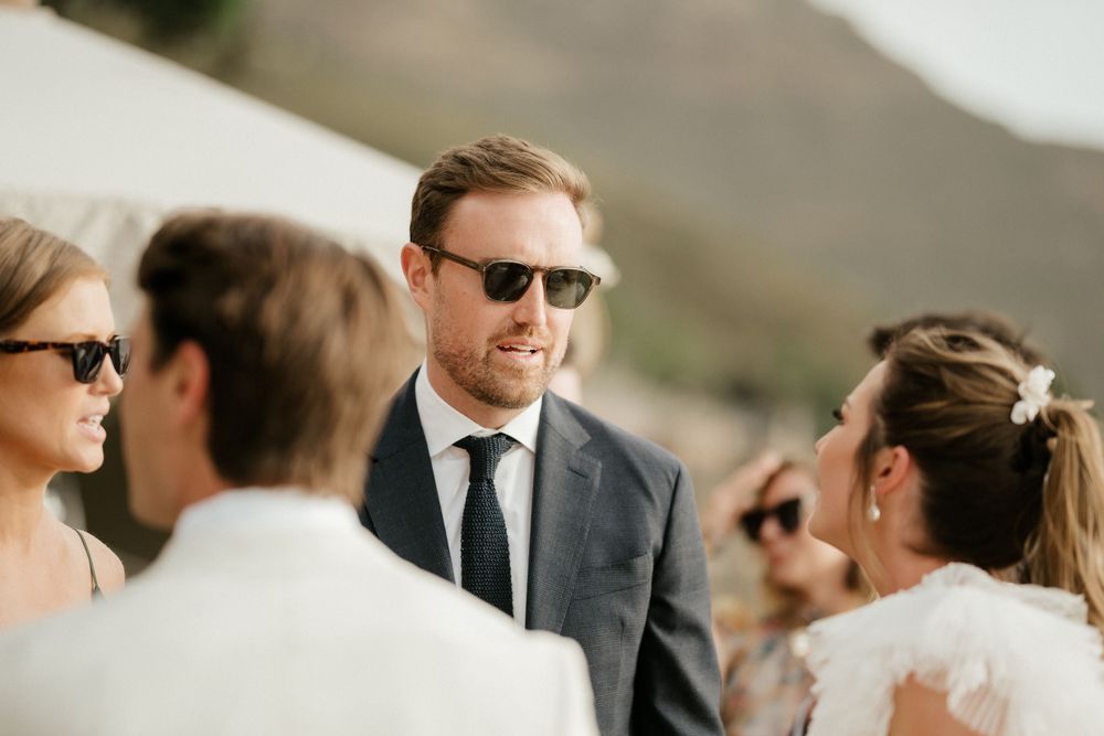 Wedding guests in formal attire gather outdoors for conversation during a sunny celebration.