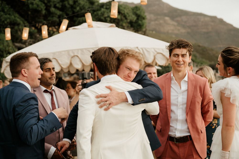 Outdoor wedding celebration with guests gathering under white tent and string lights at sunset.