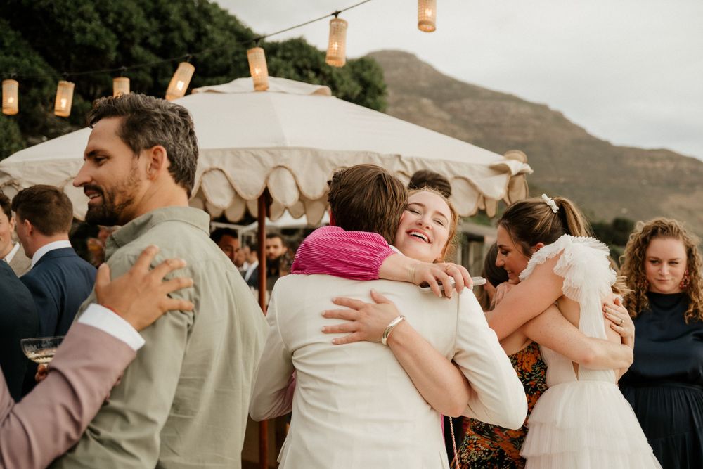 Happy guests share warm embraces during an outdoor wedding reception under white umbrellas with mountains in the background.