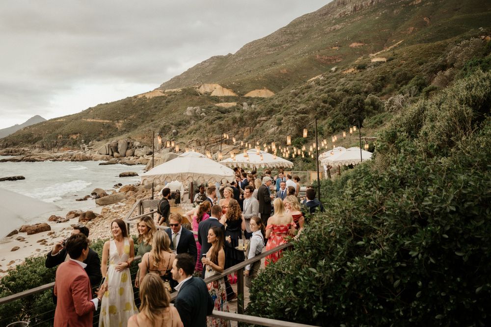 A crowd gathers along a coastal pathway for an outdoor celebration with mountain views.