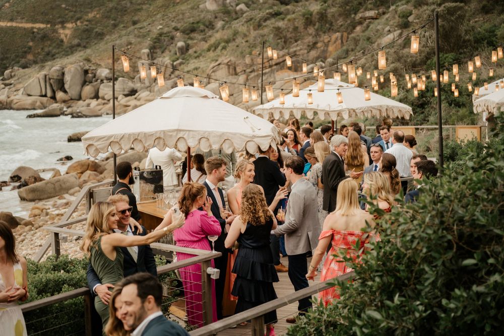 Wedding guests gather at an outdoor reception with string lights and white umbrellas along a scenic coastal venue at sunset.