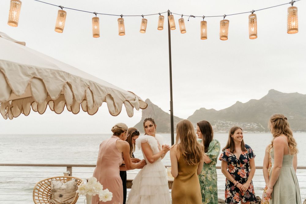 Wedding guests gather under string lights on a deck overlooking mountains and water at sunset.