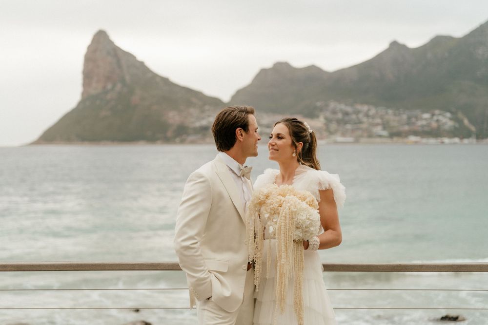 Couple in wedding attire poses on balcony overlooking scenic coastal mountains and bay.