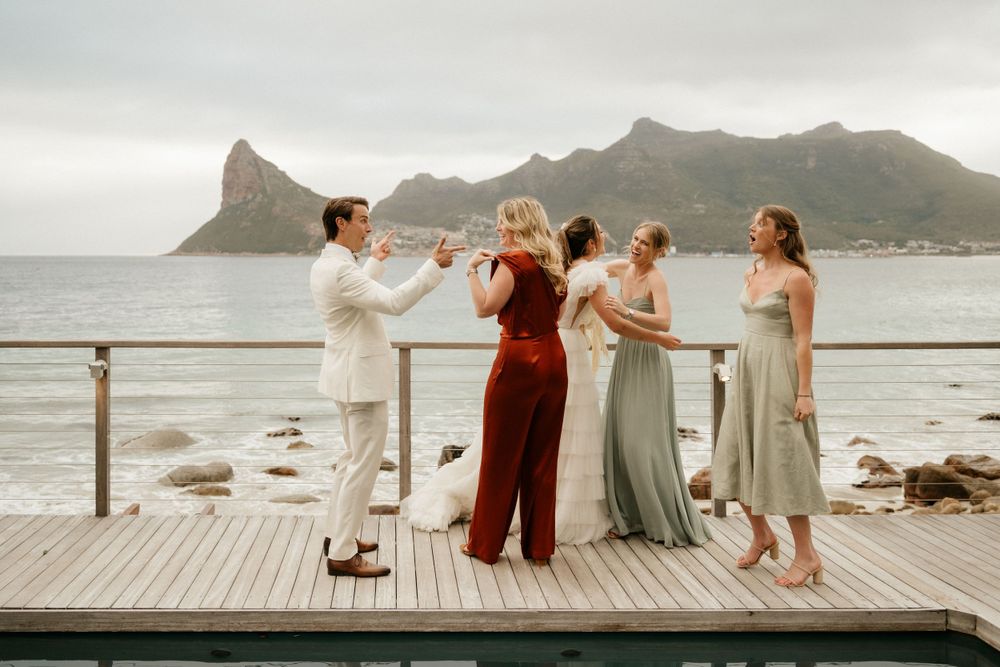 A group in elegant attire celebrates on a wooden deck overlooking a dramatic mountain coastline.