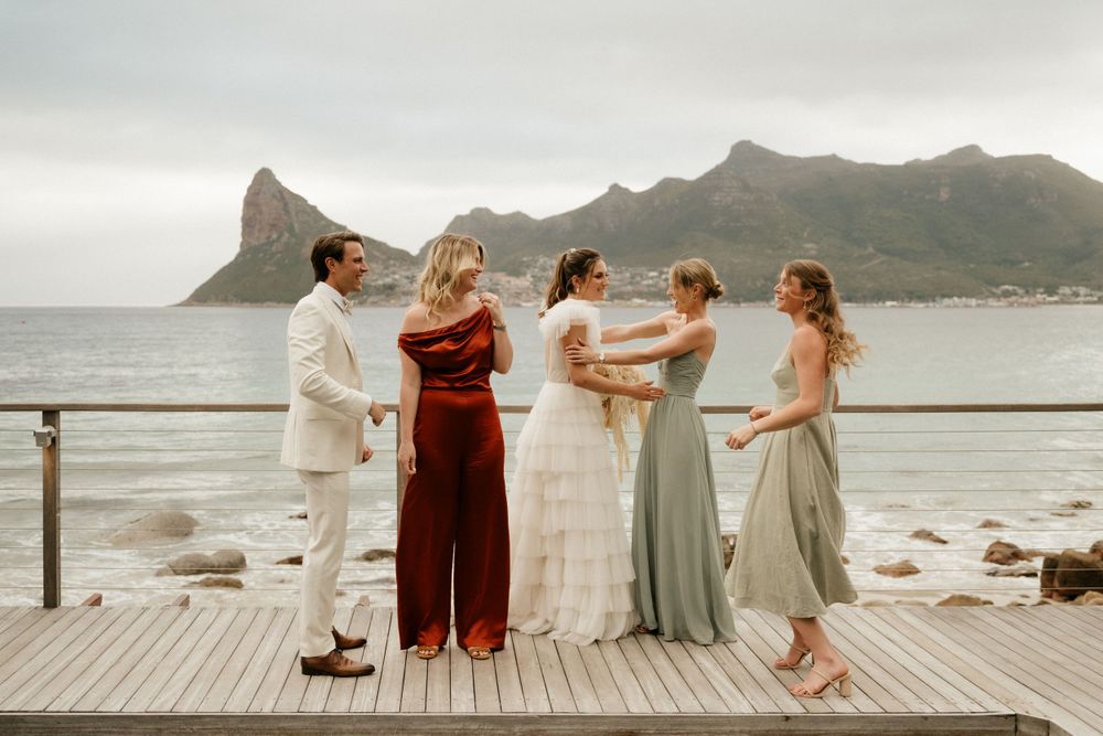 Wedding party in flowing dresses poses on wooden deck with coastal mountain backdrop.