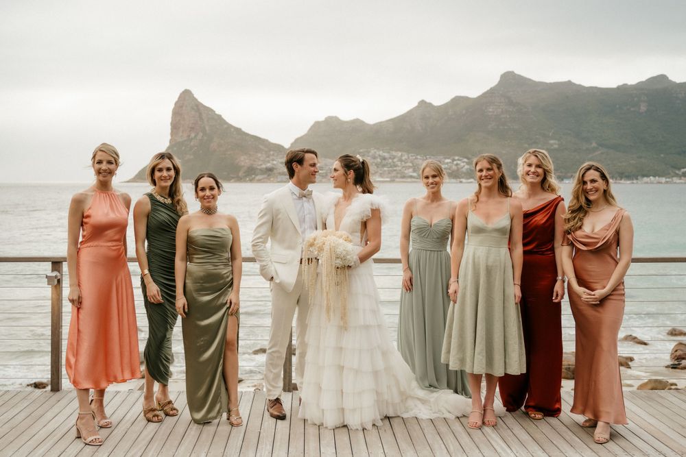 Wedding party poses on waterfront deck with dramatic mountain backdrop in mismatched formal dresses.
